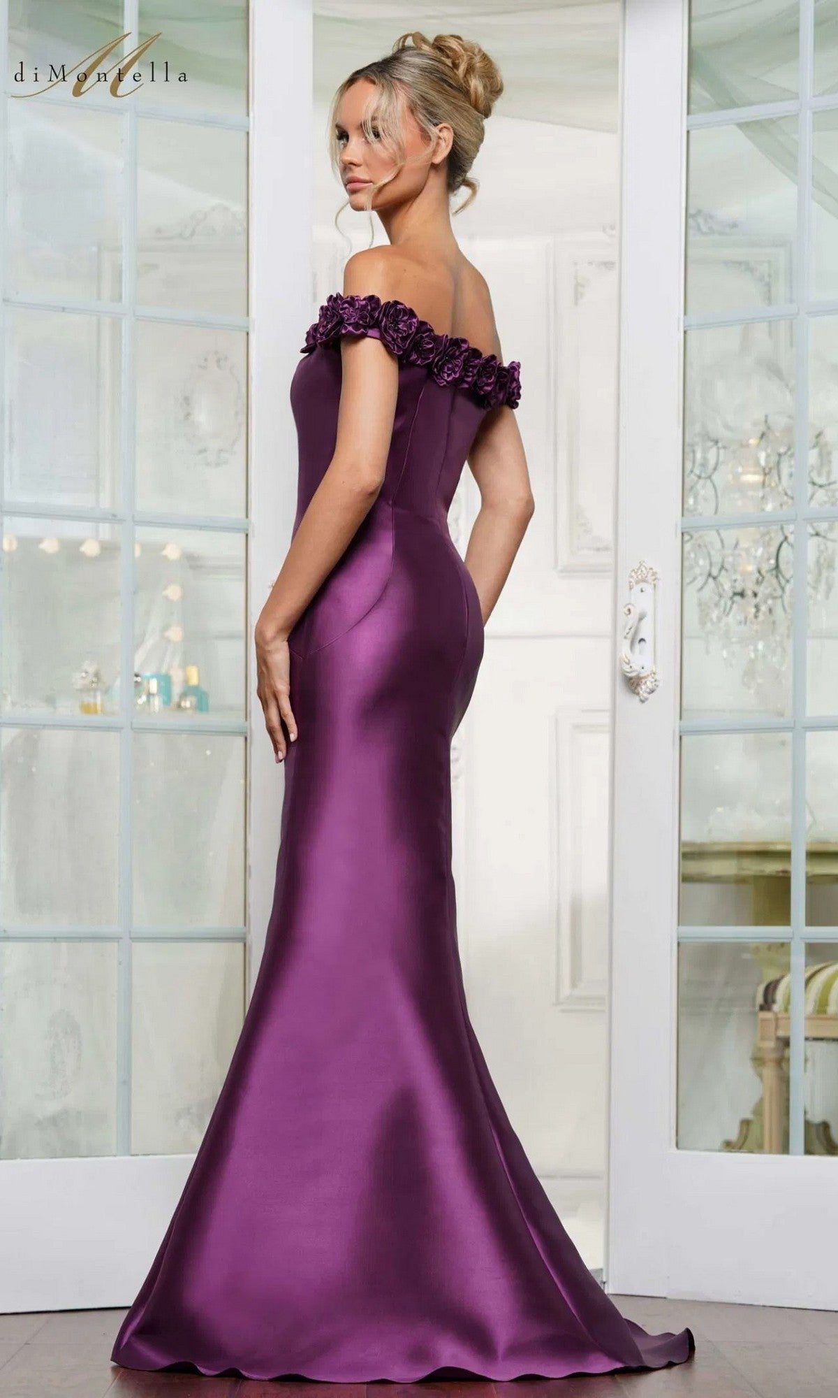 A woman in the Rina Di Montella RD3205 long formal dress, featuring off-the-shoulder purple satin, floral details, and a mermaid silhouette, stands before glass doors.
